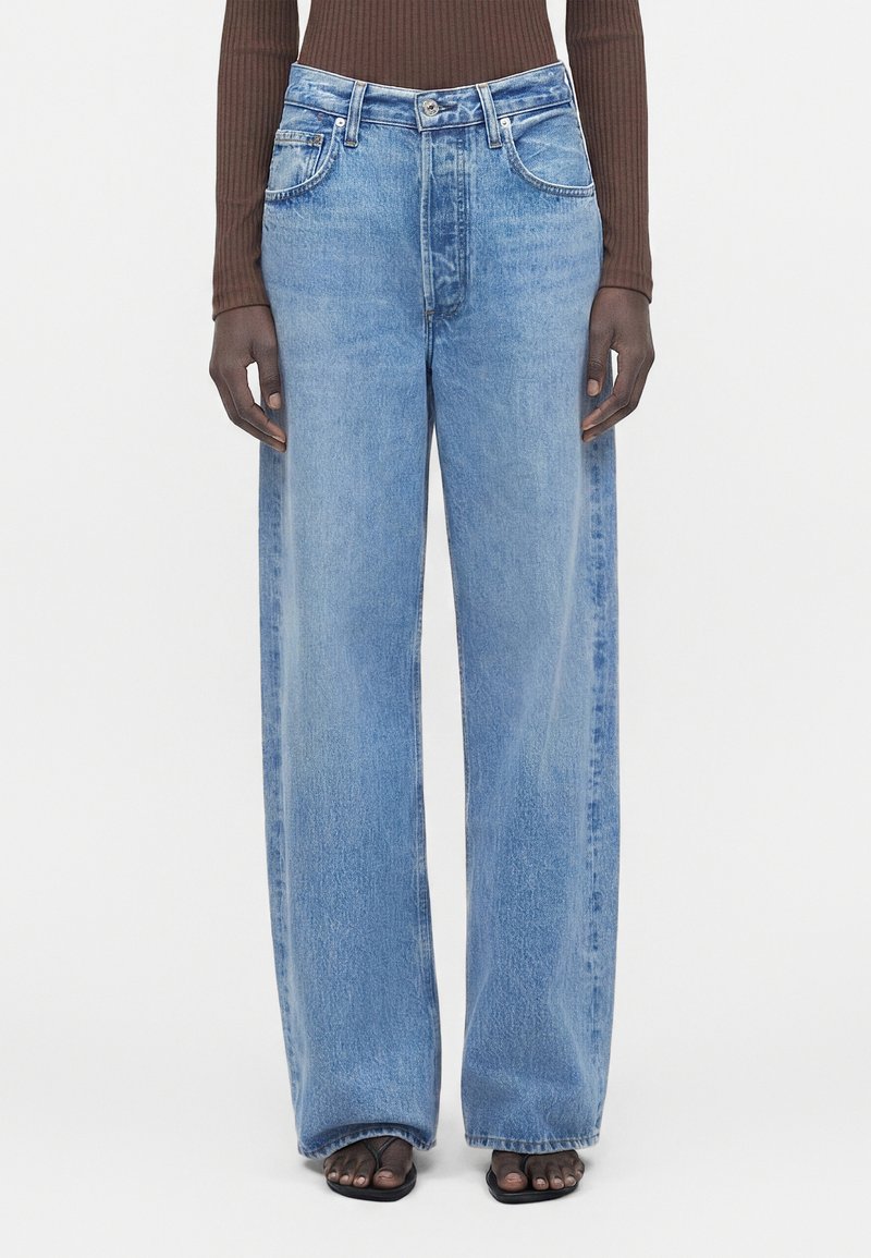 Μοντέλο με ανοιχτού μπλε τζιν παντελόνι με φαρδιά πόδια, καφέ μακρυμάνικο ριπ τοπ και μαύρα σανδάλια ανοιχτά δάχτυλα σε λευκό φόντο.