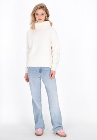 Weißer Oversized-Pullover mit Rollkragen und geripptem Saum sowie Bündchen, kombiniert mit hellblauen Straight-Leg-Jeans und rosafarbenen Sneakers.