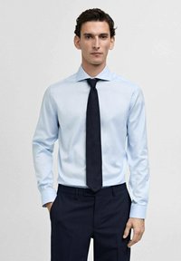 Lichtblauw overhemd met lange mouwen en een puntige kraag, gecombineerd met een donkerblauwe stropdas en donkere broek. Strakke lijnen, minimale textuur.