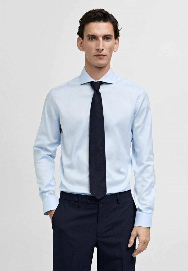 Lichtblauw overhemd met lange mouwen en een puntige kraag, gecombineerd met een donkerblauwe stropdas en donkere broek. Strakke lijnen, minimale textuur.