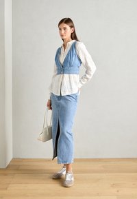 Femme se tenant à l'intérieur, portant une blouse blanche sous un gilet en jean bleu et une jupe longue en jean assortie avec des baskets beiges, tenant un sac à main blanc.