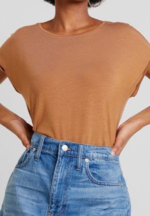 Osoba ubrana w krótką, karmelowo-brązową bluzkę wpuszczoną w wysokiej talii niebieskie dżinsy, ręce spoczywają na biodrach.