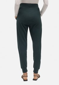Pantalones jogger verde oscuro con cintura ajustada, puños elásticos y un tejido texturizado. Combinados con sandalias planas, mostrando un diseño suave y casual.