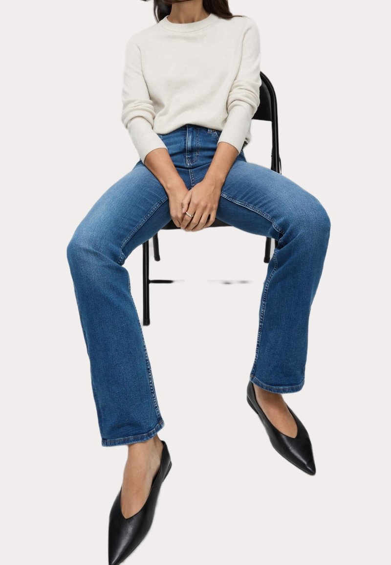 Mujer sentada en una silla negra, vistiendo un suéter blanco de manga larga, jeans azules y zapatos planos negros de punta, con las manos entrelazadas en el regazo.