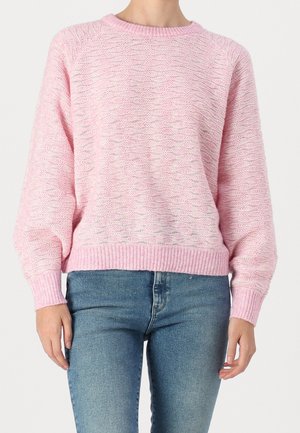 Personne portant un pull rose clair en maille lâche avec manches longues, associé à un jean bleu délavé moyen sur un fond uni.
