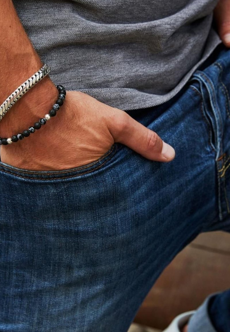 Dark blue denim jeans with a slim fit, worn with a gray fabric top. A hand rests in the pocket, showcasing a silver chain bracelet and beaded bracelet.