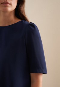 Top blu navy a maniche corte con scollatura rotonda. Il tessuto ha una consistenza liscia, caratterizzato da dettagli leggermente arricciati sulle spalle e una vestibilità aderente.