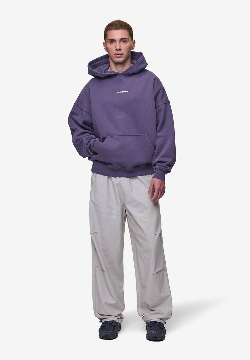 Fioletowa, oversize bluza z kapturem z przednią kieszenią i logo, zestawiona z luźnymi beżowymi spodniami i ciemnymi butami sportowymi. Gładki materiał, luźny krój.