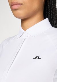 Frau, die ein weißes Hemd mit Kragen trägt, mit einem kleinen schwarzen Logo auf der linken Brust, zeigt das untere Gesicht und den Hals.
