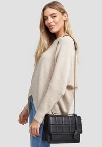 Czarna skórzana torebka z pikowanym wzorem, złotym łańcuszkiem, usztywnionym kształtem i zamknięciem na klapę. Modelka ma na sobie jasnobeżowy sweter.