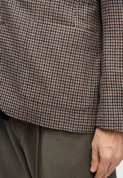 Καφέ και μαύρο σακάκι με μοτίβο houndstooth σε τσέπη πάνω σε ελαιδί πτυχωτά παντελόνια, με ορατό δεξί χέρι που ακουμπάει στο πλάι.