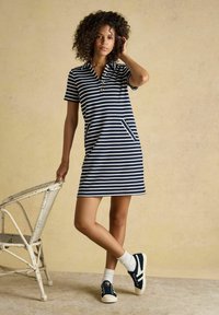 Femme en robe rayée bleu marine et blanche avec des boutons, chaussettes blanches et baskets, debout à côté d'une chaise en osier blanche sur fond beige.