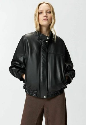 Giovane con capelli chiari indossa una giacca bomber nera in pelle e pantaloni marroni, in piedi con le mani nelle tasche della giacca davanti a uno sfondo neutro.