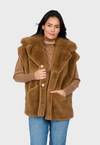 Gilet in finta pelliccia marrone con colletto ampio, chiusura con bottone dorato e tasche frontali, indossato sopra un maglione beige e jeans blu. Texture morbida.