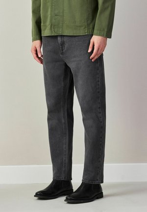 Mężczyzna noszący czarne buty Chelsea, wyblakłe czarne dżinsy i oliwkowo zieloną kurtkę, stojący wewnątrz pomieszczenia na tle jasnoszarej ściany.