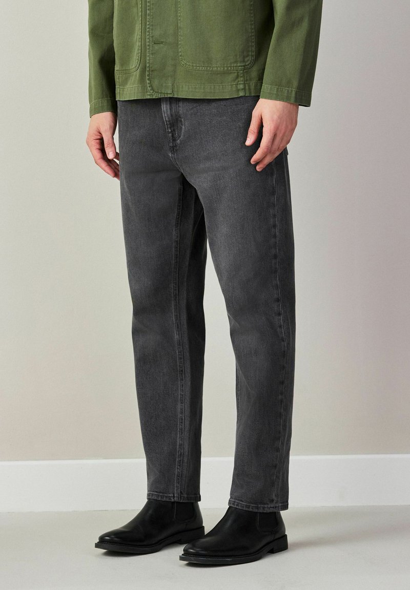 Hombre con botas Chelsea negras, jeans negros desgastados y una chaqueta verde oliva, de pie en interiores contra una pared gris clara.