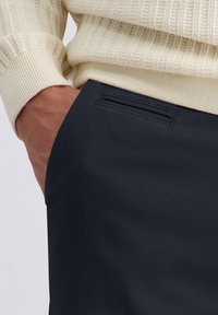 Marineblauwe broek met een gestructureerde afwerking, voorzien van een klein horizontaal zakje aan de voorkant en gecombineerd met een crèmekleurige ribbeltrui.