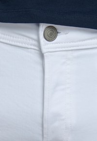 Detailní záběr na pásku bílých džín s kovovým knoflíkem a tmavomodrou tkaninou nahoře.