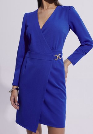 Kobieta ubrana w królewsko niebieską sukienkę kopertową z długim rękawem, z ozdobnym złotym klamrą w talii, pozująca z jedną ręką na biodrze, a drugą swobodnie opadającą.