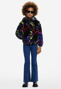 Tekoturkistakki, jossa värikkäitä kuvioita neonpunaisena, vihreänä ja sinisenä. Yhdistetty liehuhousuihin ja mustiin nilkkureihin.