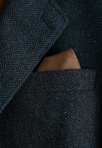 Veste bleu foncé à texture chevrons avec un mouchoir de poche en satin marron glissé dans la poche poitrine.