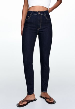 Jean slim bleu foncé avec surpiqûres contrastantes, porté avec des tongs noires et un top court blanc, la zone médiane visible.