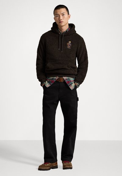 Bruine fleece hoodie met een trekkoordcapuchon, een geruite shirt eronder, zwarte broek en bruine schoenen. Heeft een geborduurd logo op de borst.