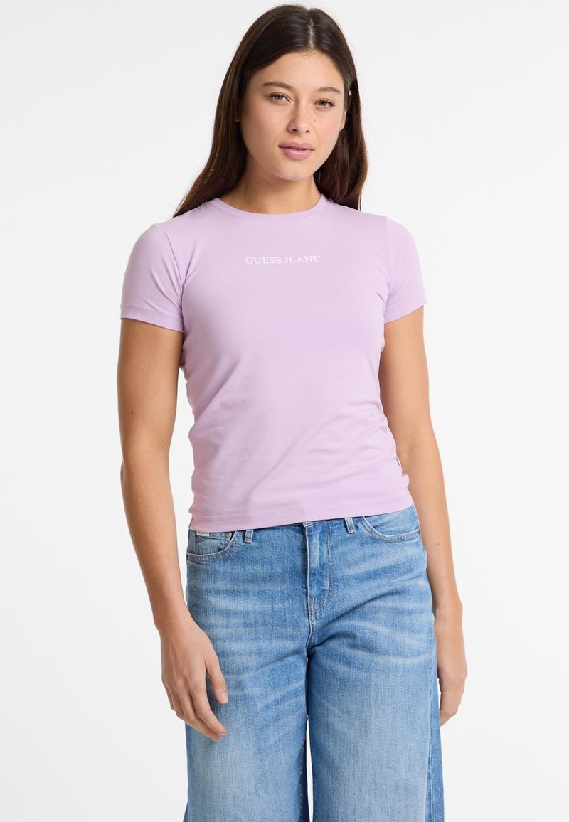 Lila aansluitend t-shirt met korte mouwen, logo "GUESS JEANS" in het wit op de borst, gedragen met lichte blauwe wijde jeans.