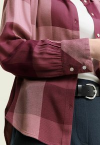 Chemise à carreaux bordeaux et rose clair avec des poignets boutonnés, ourlet arrondi et texture douce, portée sur un haut blanc et associée à une ceinture noire.