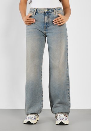 Person iført løstsiddende, lyseblå jeans og hvide sneakers, der står med hænderne nær forlommerne mod en ensfarvet baggrund.