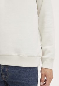 Witte sweatshirt met geribbelde manchetten en zoom, gemaakt van zachte stof. Zichtbaar merketiket aan de linkerkant. Draag over donkere jeans.