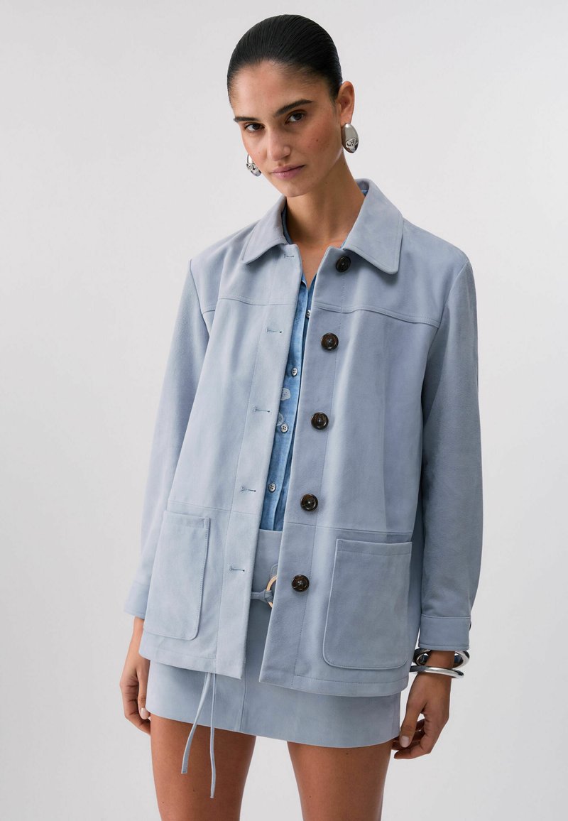 Mujer con chaqueta de ante azul claro con grandes botones, combinada con una falda a juego y camisa de mezclilla, complementada con pendientes y pulseras plateadas.