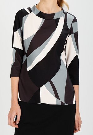 Femme portant un haut à manches longues ajusté avec des motifs géométriques abstraits en noir, blanc et gris, associé à une jupe noire.