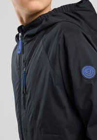 Musta kevyt hupparitakki, jossa on elastiset hihansuut, varustettuna vetoketjullisella rintataskulla, sinisillä yksityiskohdilla ja pyöreällä logolla hihassa.