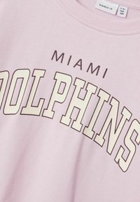 Gaiši rozā džemperis ar uzrakstu "MIAMI DOLPHINS" krēmkrāsā un brūnā tonī. Ribēta apkakle un mīksta tekstūra nodrošina komfortu.