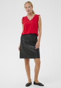 Crvena majica bez rukava s čipkastim rubom, uparena s crnom kožnom suknjom do koljena. Model nosi crne ravne cipele. Jednostavan, minimalistički stil.