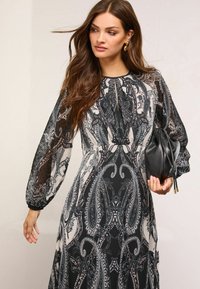 Rochie alb-negru cu model paisley, mâneci lungi transparente, corset plisat și fustă vaporoasă. Asortată cu o geantă neagră.