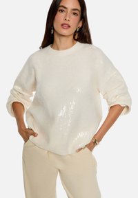 Pull en maille blanche avec une texture douce, un col rond, et orné de paillettes sur un côté. Associé à un pantalon beige clair.