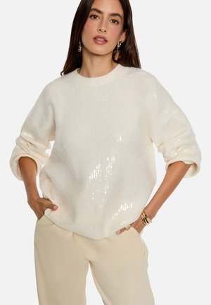Pull en maille blanche avec une texture douce, un col rond, et orné de paillettes sur un côté. Associé à un pantalon beige clair.