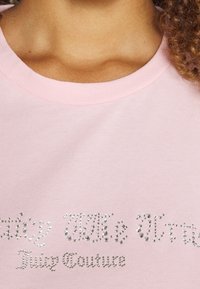 Růžové bavlněné tričko s kulatým výstřihem, ozdobené stříbrnými kamínky uspořádanými do stylizovaného textu "Juicy Couture."