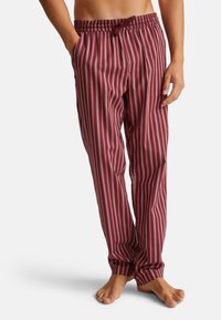 Man in een rood en wit verticaal gestreepte pyjamabroek met zijn handen in zijn zakken, staande op blote voeten voor een witte achtergrond.