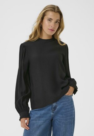 Chemisier noir à manches longues avec épaulettes bouffantes et poignets à volants, fabriqué dans un tissu lisse. Porté avec un jean bleu clair, offrant une coupe décontractée.