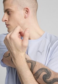 Lyseblå atletisk t-shirt med korte ærmer, der har et lille gråt Puma-logo. Nærbillede af en persons arm med en tatovering, som flexer hånden.