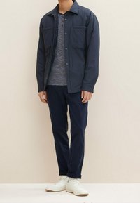 Námořnická lehká bunda s dvěma náprsními kapsami přes šedé tričko henley, v kombinaci s tmavě námořnickými kalhotami a bílými teniskami.