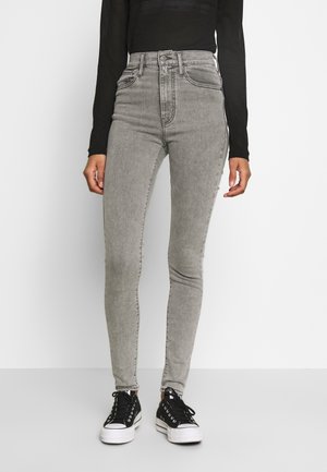 Kvinde iført højtaljede grå skinny jeans, sort langærmet top og sorte lave sneakers, stående mod en hvid baggrund.