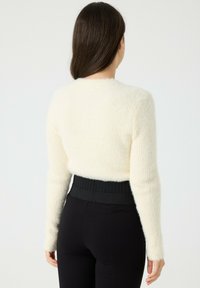 Mujer con cabello largo castaño, vestida con un suéter blanco de felpa y pantalones negros de cintura alta, de pie de espaldas contra un fondo sencillo.