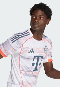 Biały sportowy trykot z delikatnymi różowymi i szarymi falistymi wzorami, z emblematem i logo. Krótkie rękawy z pomarańczowymi wykończeniami i czarnymi paskami.