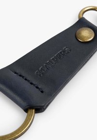 Llave de cuero negro con herrajes de latón, que presenta el texto "SCALPERS" grabado y costuras decorativas a lo largo de los bordes.