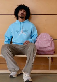 Φωτεινή γαλάζια μπλούζα με κουκούλα και λευκό σχέδιο, γκρι ριγέ παντελόνι, κρέμ αθλητικά παπούτσια και ροζ σακίδιο πλάτης με σχέδια, τοποθετημένα σε ένα ξύλινο παγκάκι.