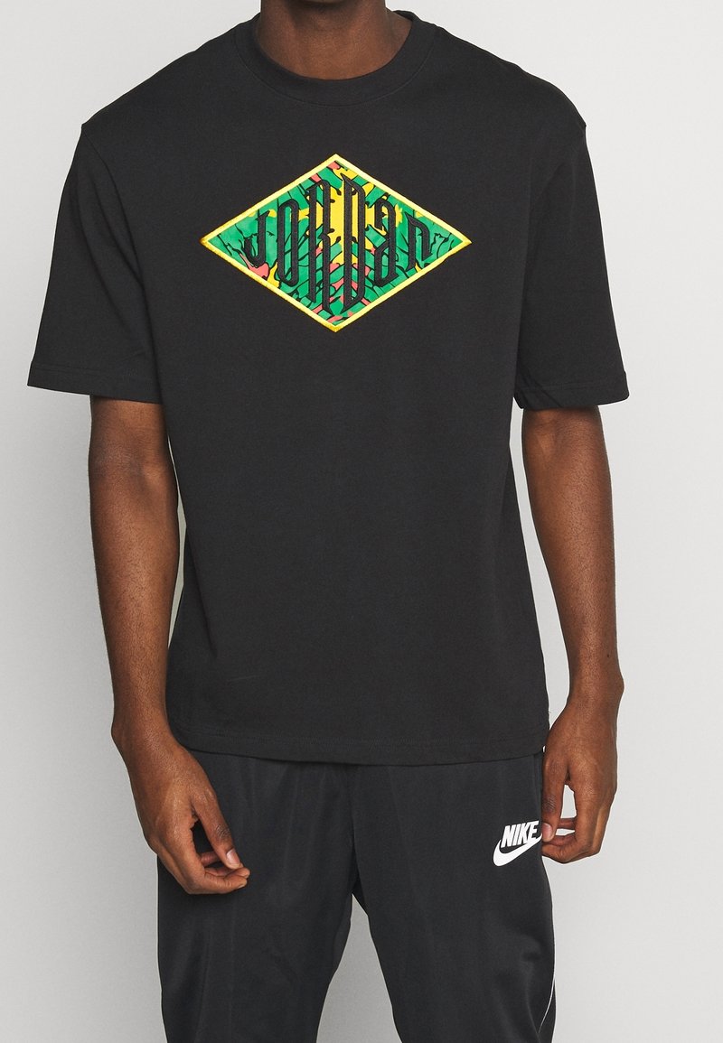 Homme portant un t-shirt noir avec un motif coloré en forme de diamant et un pantalon de survêtement Nike noir avec un logo blanc sur la cuisse gauche.